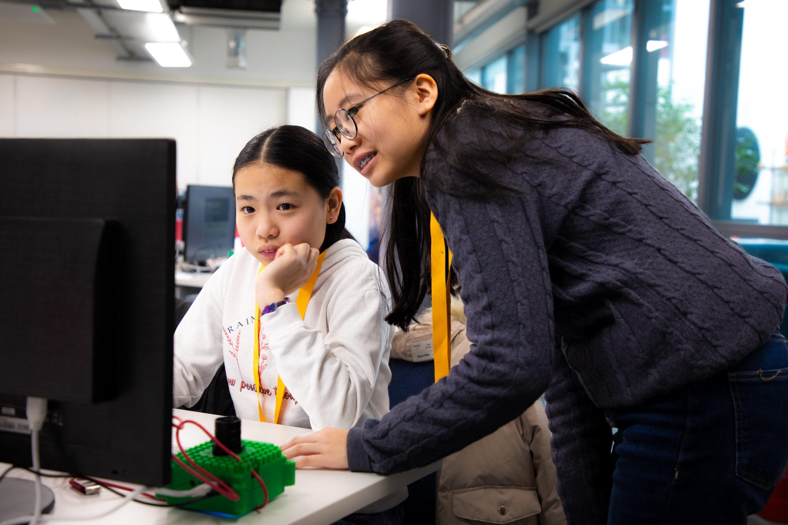 Two students work together at a computer during a coding activity. One student is seated at the workstation while another leans over to view the screen, with electronic components and programming equipment visible on the desk in a classroom or workshop environment.